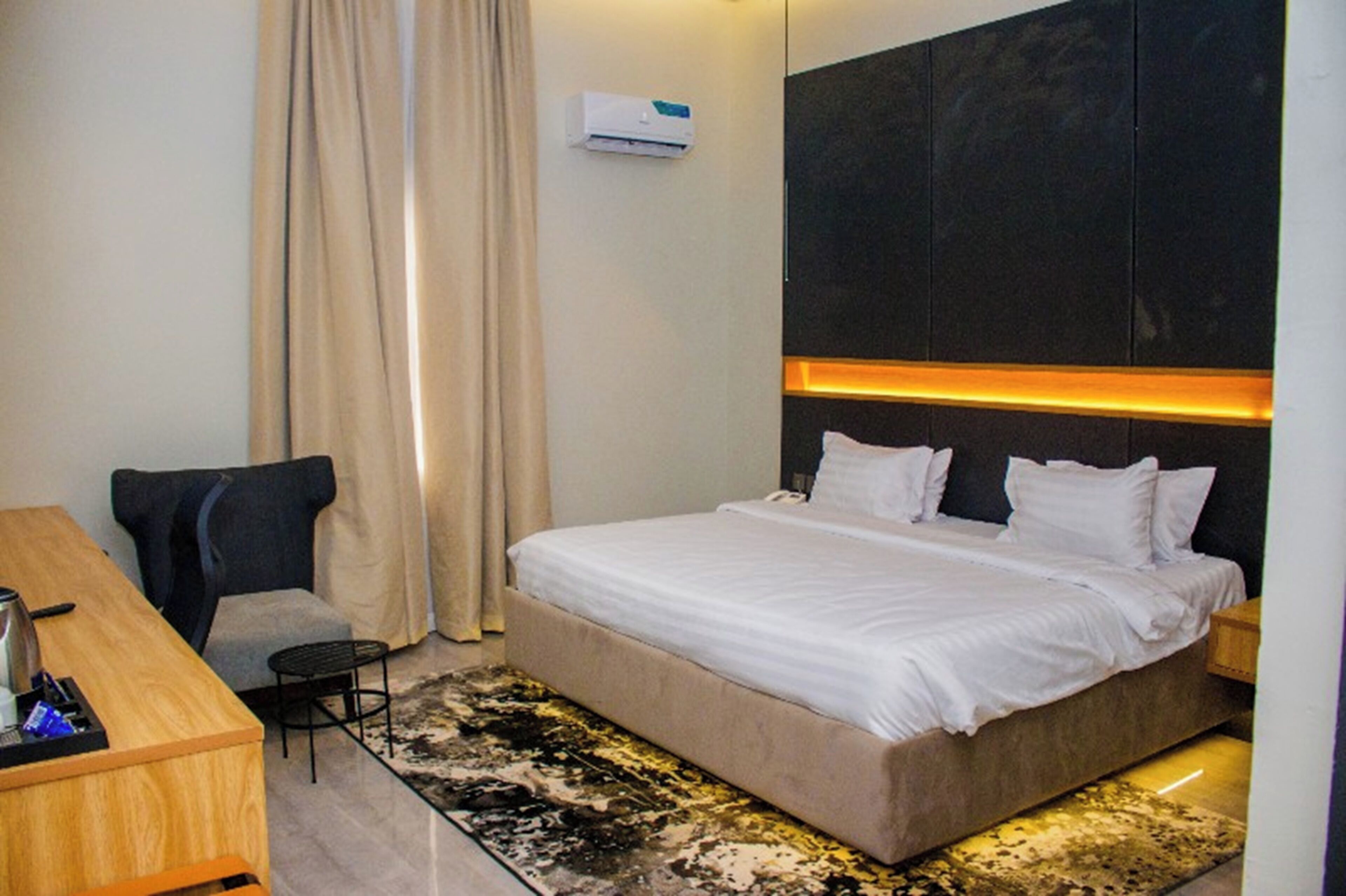 A hotel room with a bed, a desk, a chair, an air conditioning unit, and a patterned rug.