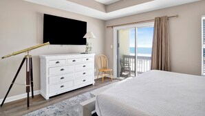 A bedroom with a large bed, a TV mounted on the wall, a wooden chair, a white dresser, and a balcony with curtains. - Gulf Front Duplex | Shared Pool | West Beach (Gulf Shores)