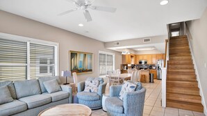 A spacious living room with a sofa, armchairs, and a coffee table. A kitchen is visible in the background with wooden cabinets and a staircase. - Gulf Front Duplex | Shared Pool | West Beach (Gulf Shores)