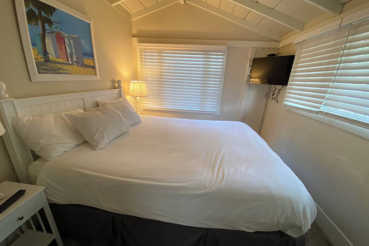 Une chambre à coucher avec un lit, des rideaux blancs, un téléviseur fixé au mur et un tableau accroché au mur.