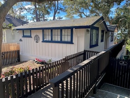 A small, single-story building with a dark blue trim and white siding, a wooden fence, and a small garden area with pink flowers.