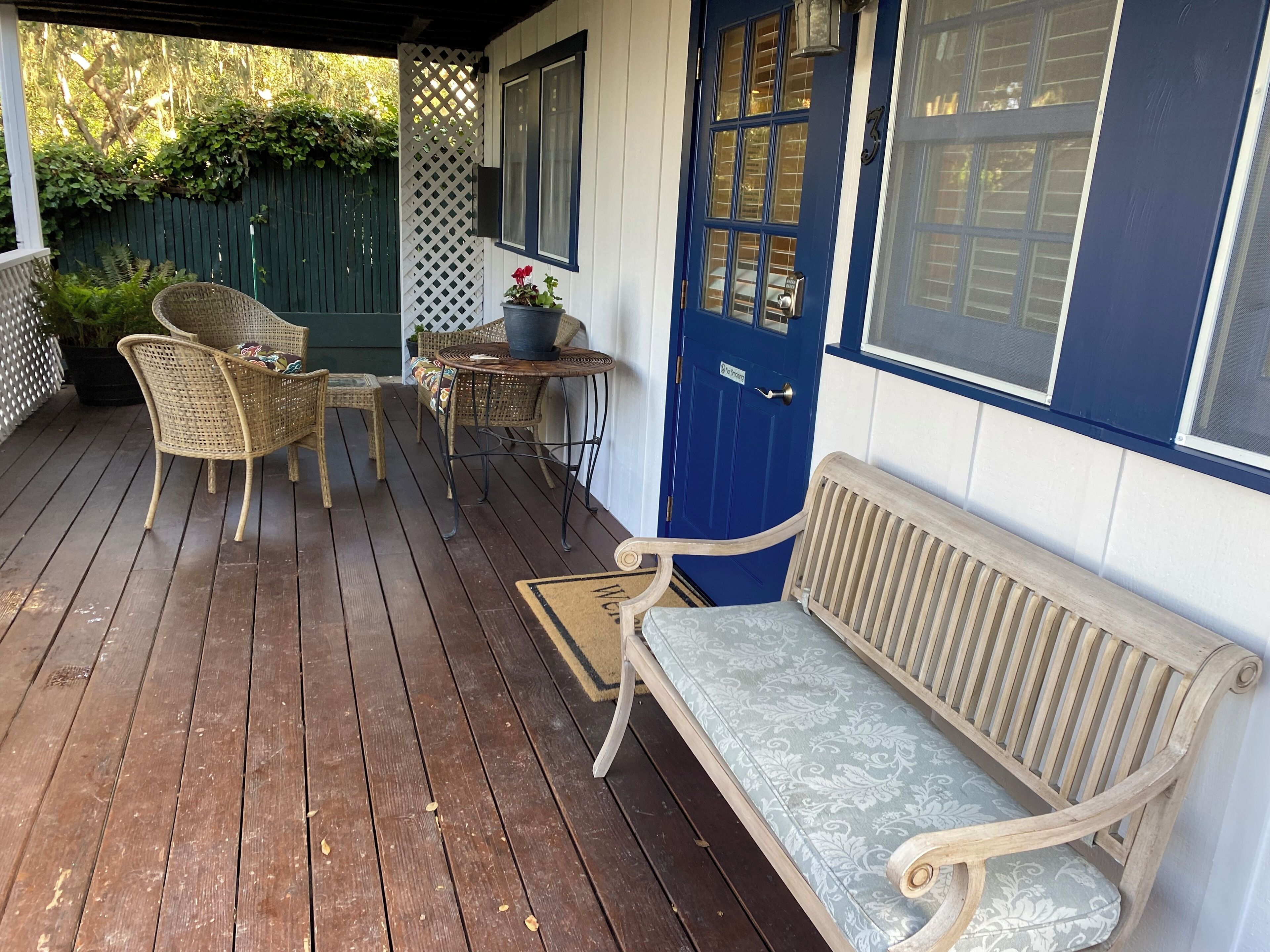 Eine Holzterrasse mit Korbstühlen und einer Bank, eine blaue Tür und ein kleiner Tisch mit einem Blumentopf.