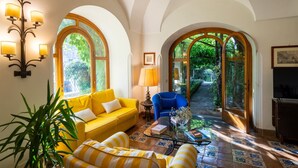 A bright living room with a yellow sofa, a blue armchair, and a glass coffee table. There are large arched windows and a lamp on the wall. - Villa Maelle by My Villa Collection (Positano)