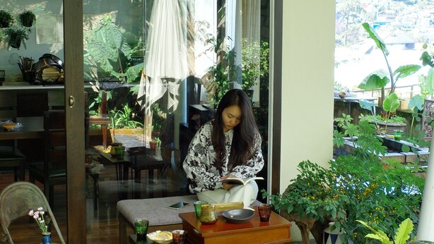 A woman sitting at a table in a room with a tiled floor, surrounded by potted plants and a glass door leading to an outdoor area. - Pha Boutique House (Xuan Huong - Da Lat)