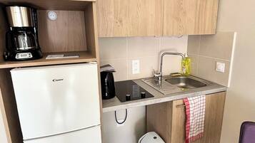 Eine kleine Küche mit einem weißen Kühlschrank, einer Kaffeemaschine, einem Spülbecken und einem Mülleimer.