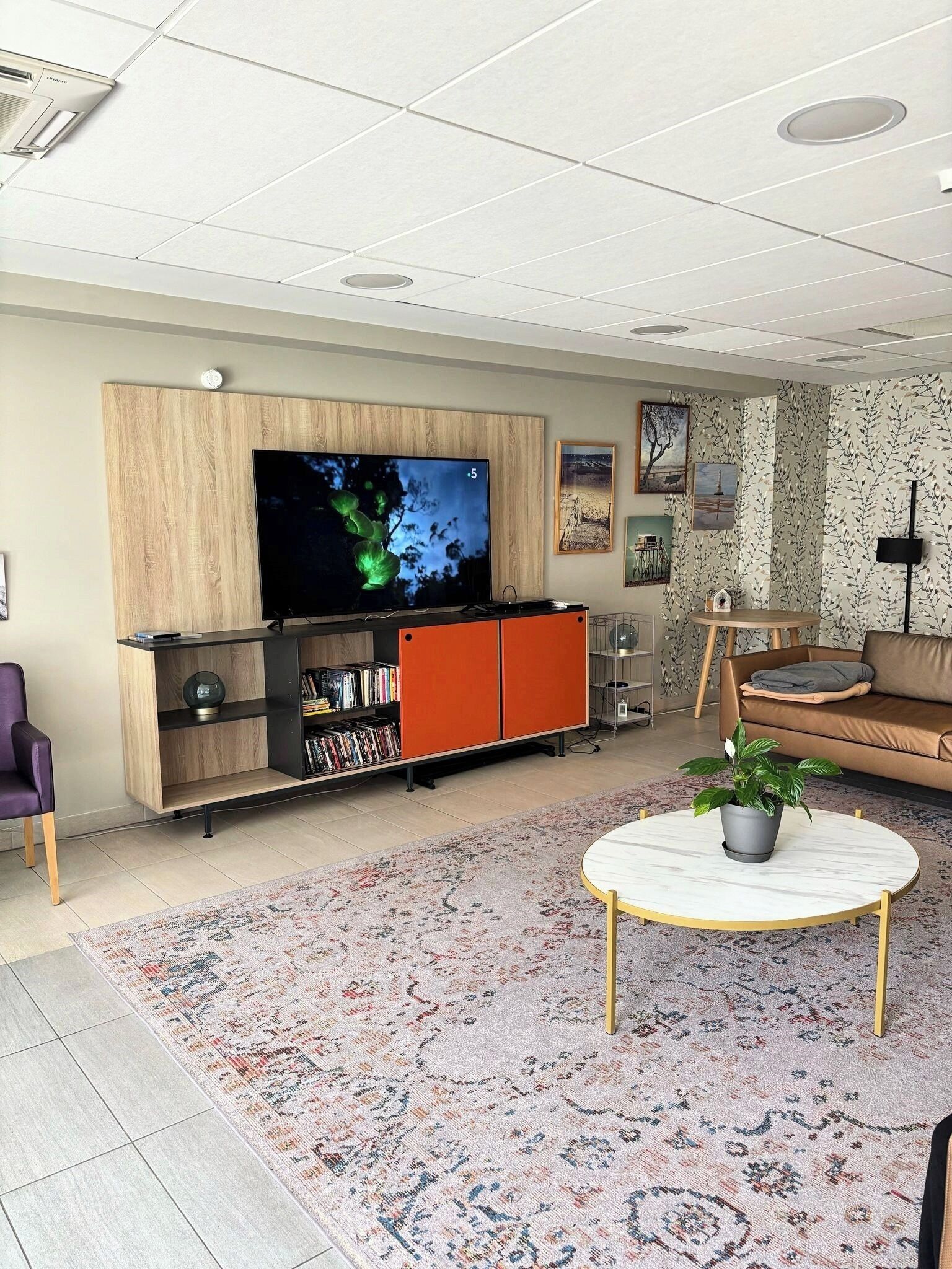 Una sala de estar con televisor de pantalla plana, armarios de madera y color naranja, una mesa redonda de centro con una planta, y una alfombra estampada.