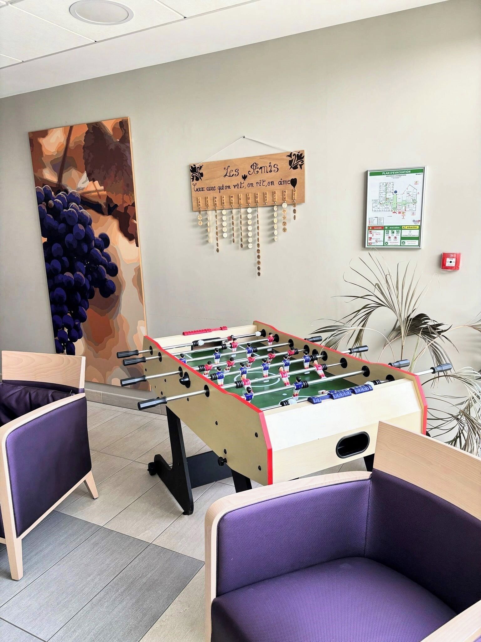 Una stanza con un tavolo da calcio, due poltrone viola, un'opera d'arte raffigurante grappoli d'uva, un cartello di legno con testo in francese e un rilevatore di fumo.