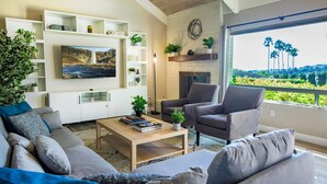 Een moderne woonkamer met een bank, fauteuils, een salontafel en een open haard. Er zijn planken met decoratieve objecten en een aan de muur bevestigde televisie.