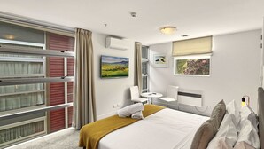 A hotel room with a bed, a small table, chairs, a window with curtains, and artwork on the walls.