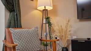 Een moderne woonkamer met een houten fauteuil, een kussen met een patroon, een vloerlamp, een potplant en een hoge vaas met pluisgras.