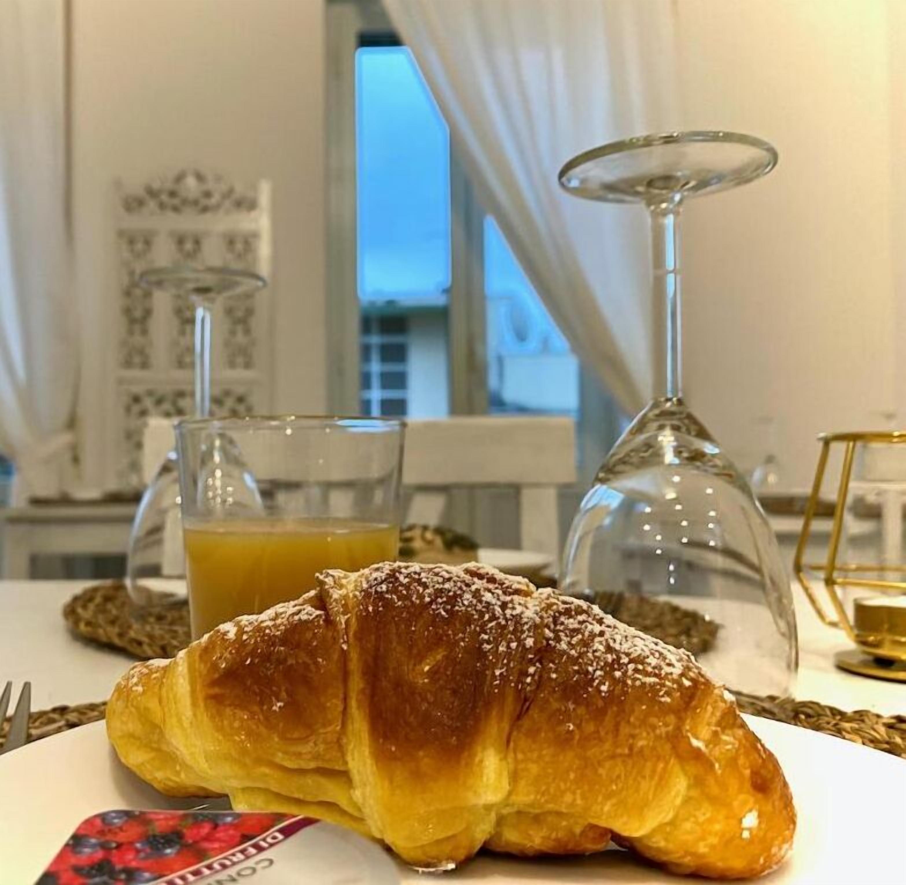 A croissant on a plate with a glass of orange juice and a wine glass in the background.