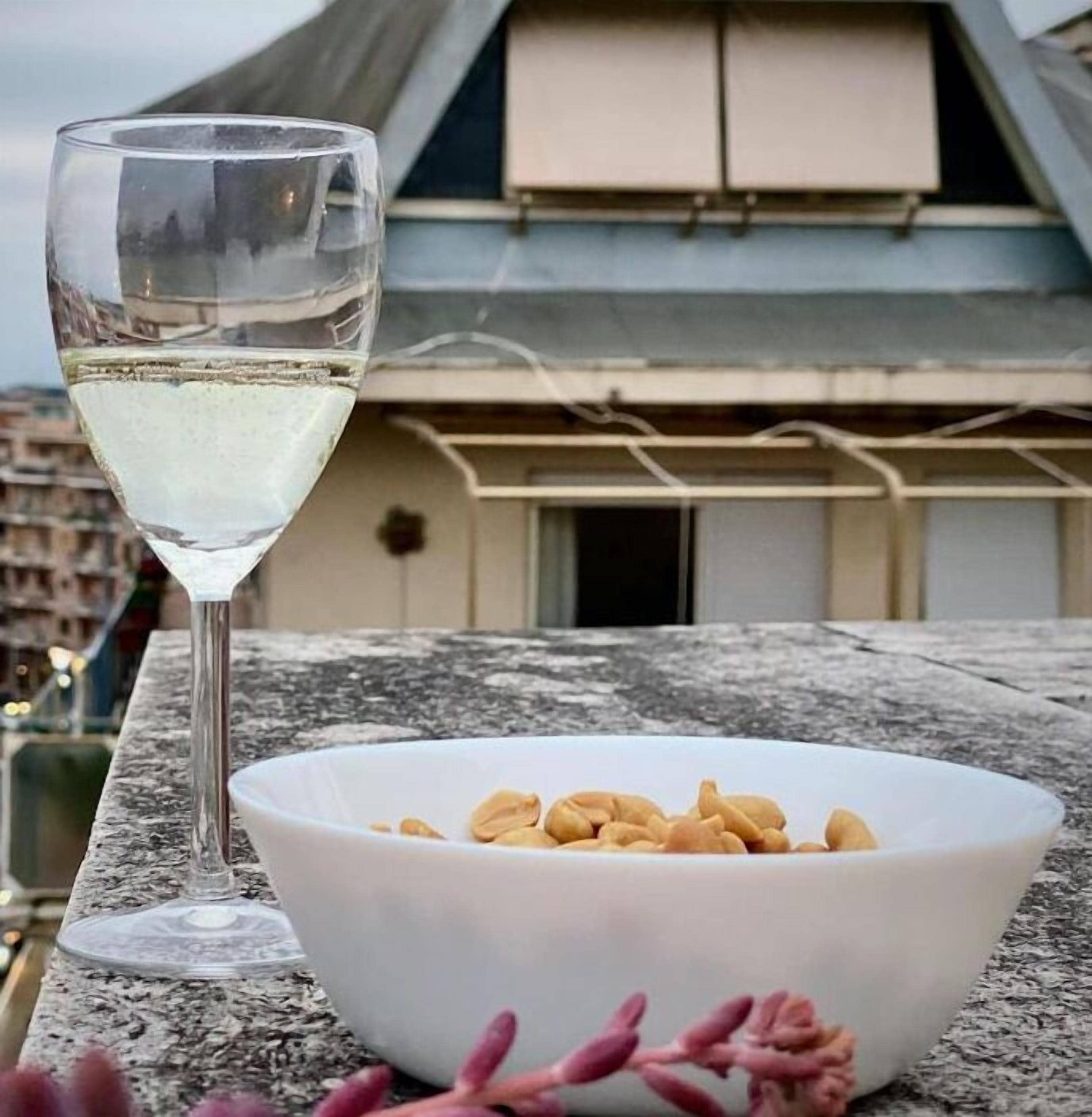 A glass of white wine on a table with a bowl of peanuts and some pink flowers.