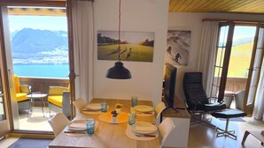 Un coin repas avec une table en bois dressée pour quatre personnes, entourée de chaises blanches. Une grande fenêtre offre une vue sur les montagnes et un plan d’eau.