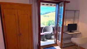 Une chambre avec une grande armoire en bois, un lit recouvert d’une couverture rose, un bureau avec un ordinateur, une chaise et une vue sur un balcon aménagé avec des meubles en osier.
