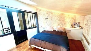 A bedroom with a stone wall, a bed with a dark cover, a wooden floor, and a window with a black frame. - Exceptional house in Nieul-sur-Mer (Nieul-sur-Mer)
