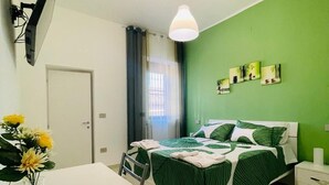 A bedroom with a bed, a desk, and a chair. There is a window with curtains, a wall-mounted TV, and a ceiling light. - Guest House " La Corte" Green Orchid. An oasis of serenity and refinement. (Zungoli)