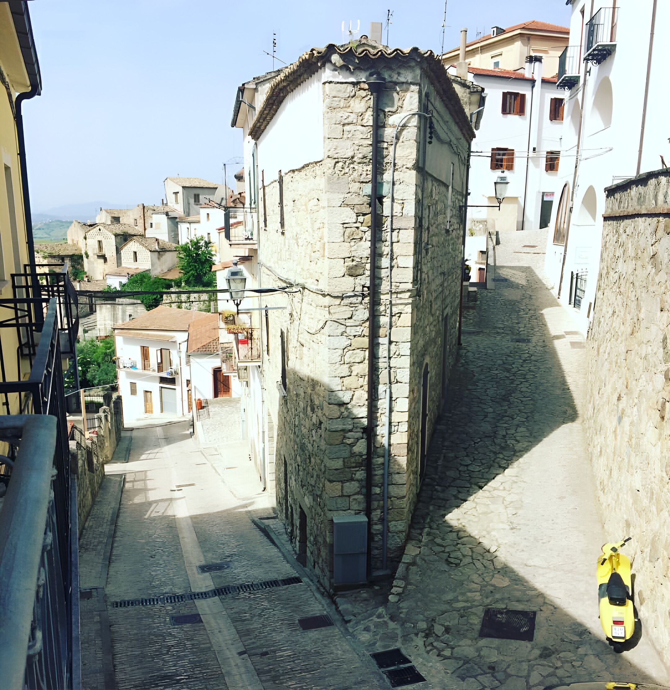 Una estrecha calle empedrada flanqueada por edificios de piedra con arquitectura tradicional, una moto estacionada a la derecha, y un cielo despejado.