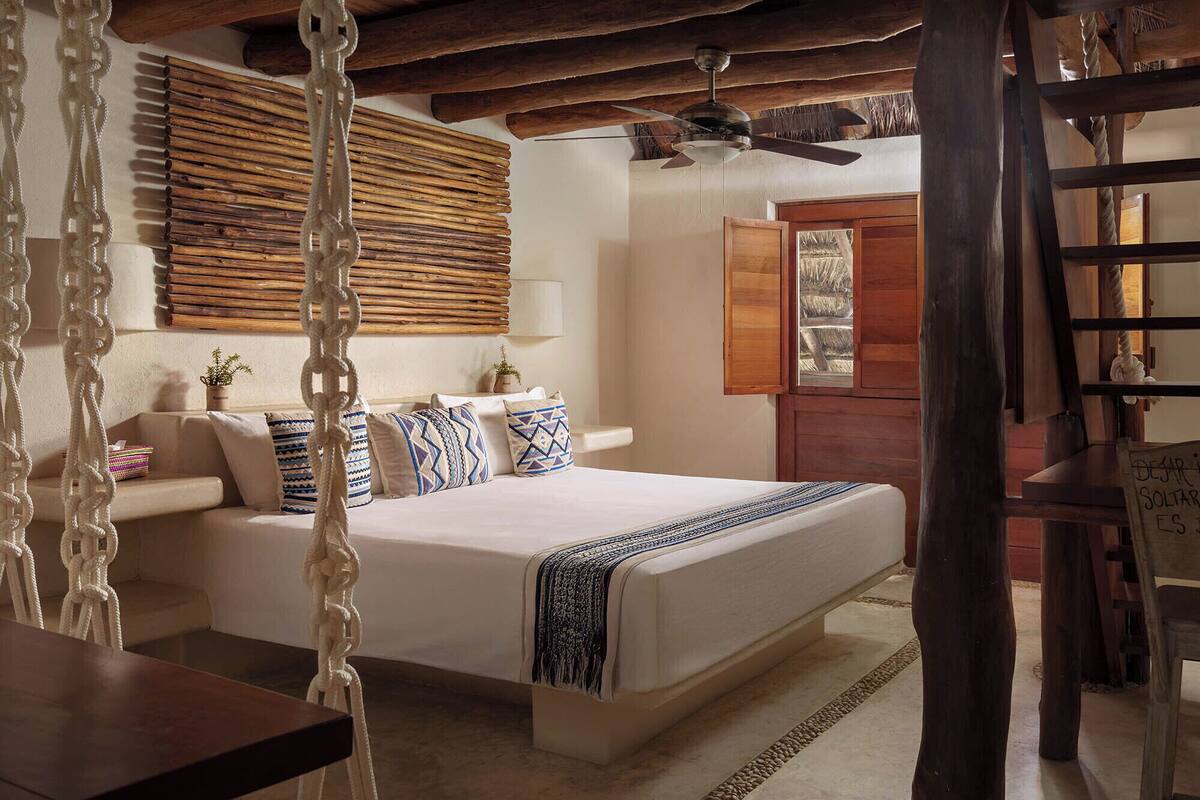 Une chambre confortable avec un lit, un fauteuil suspendu, un plafond en bois et une fenêtre avec des stores.