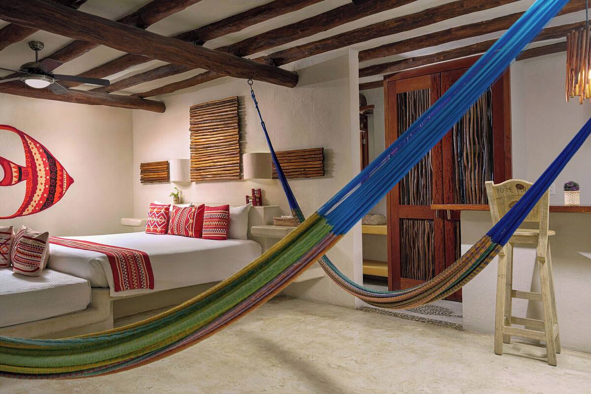 Une chambre avec un hamac, un lit agrémenté d’oreillers à motifs, des poutres en bois et un ventilateur de plafond.