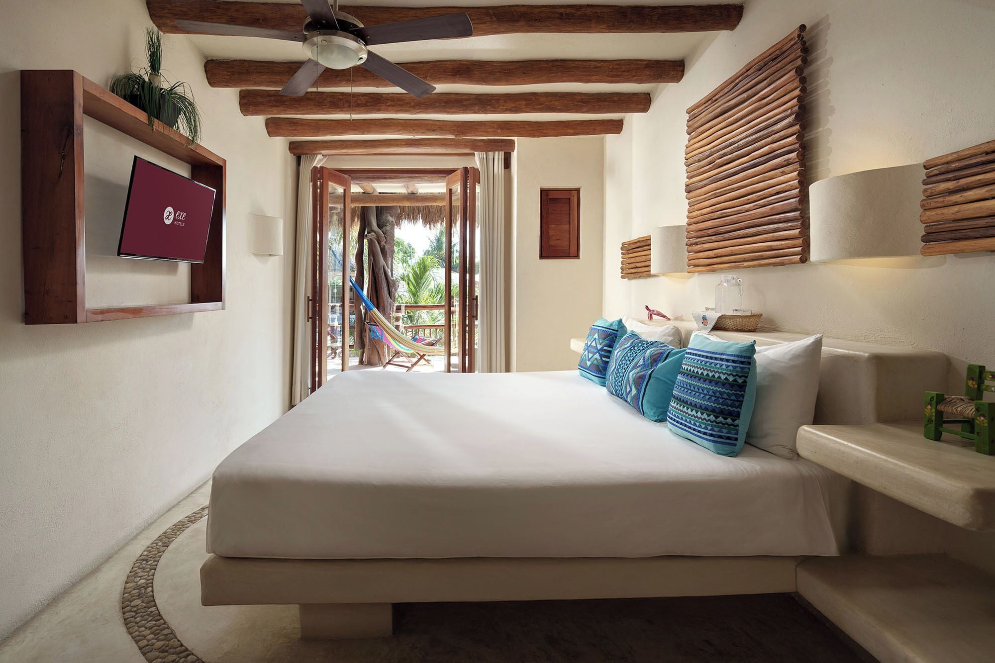 A bedroom with a large bed, a ceiling fan, wooden beams, a TV, and a view of the outdoors.