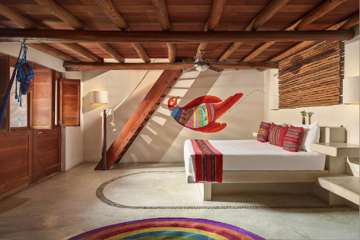 Un dormitorio con techo de madera, una hamaca, una cama con almohadones coloridos y una alfombra con diseño de arcoíris.