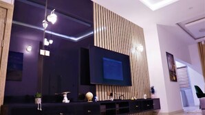 Ein modernes Wohnzimmer mit einem großen Flachbildfernseher, der an einer Wand aus Holzpaneelen befestigt ist, einer dunklen Unterhaltungseinheit darunter und einer Deckenleuchte.