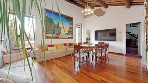 Amplia sala de estar con techo de madera, un cuadro grande, una mesa de comedor con sillas, un sofá y un televisor montado en la pared.