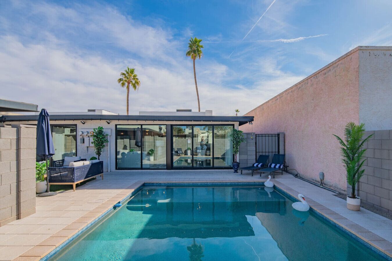 A modern outdoor pool area with a glass-enclosed patio, lounge chairs, and palm trees.