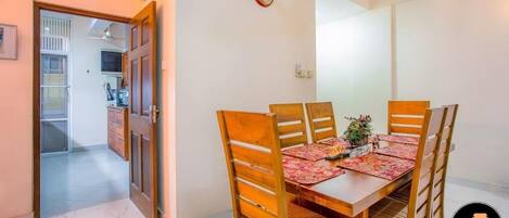 A dining area with a wooden table and chairs, a clock on the wall, and a kitchen in the background.