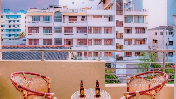 Un balcón con sillas de mimbre y una mesa pequeña, con vistas a un paisaje urbano con edificios y cielo despejado.