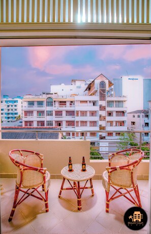 A balcony with wicker chairs and a small table, overlooking a cityscape with buildings and a clear sky.