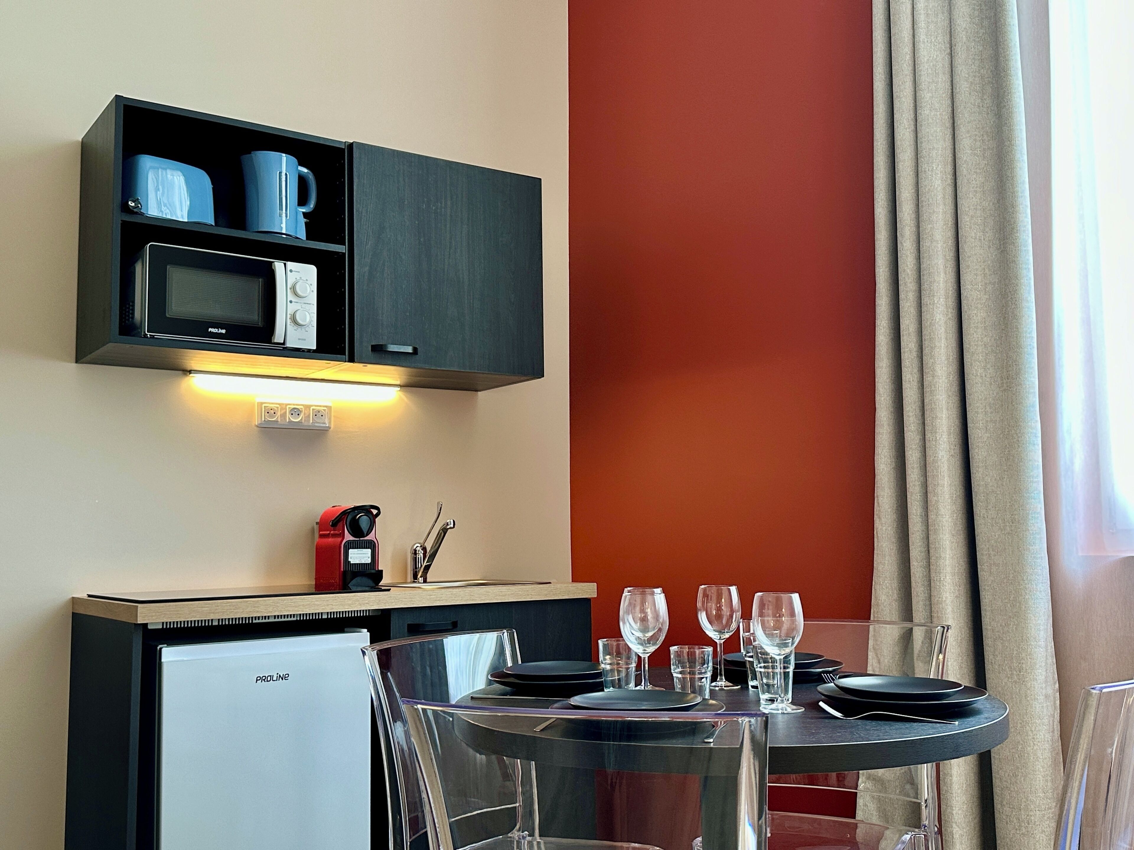 Une cuisine compacte équipée d’un four à micro-ondes, d’une bouilloire et d’une machine à café, un coin repas avec une table pour deux personnes, et un mur d’accent de couleur rouge.