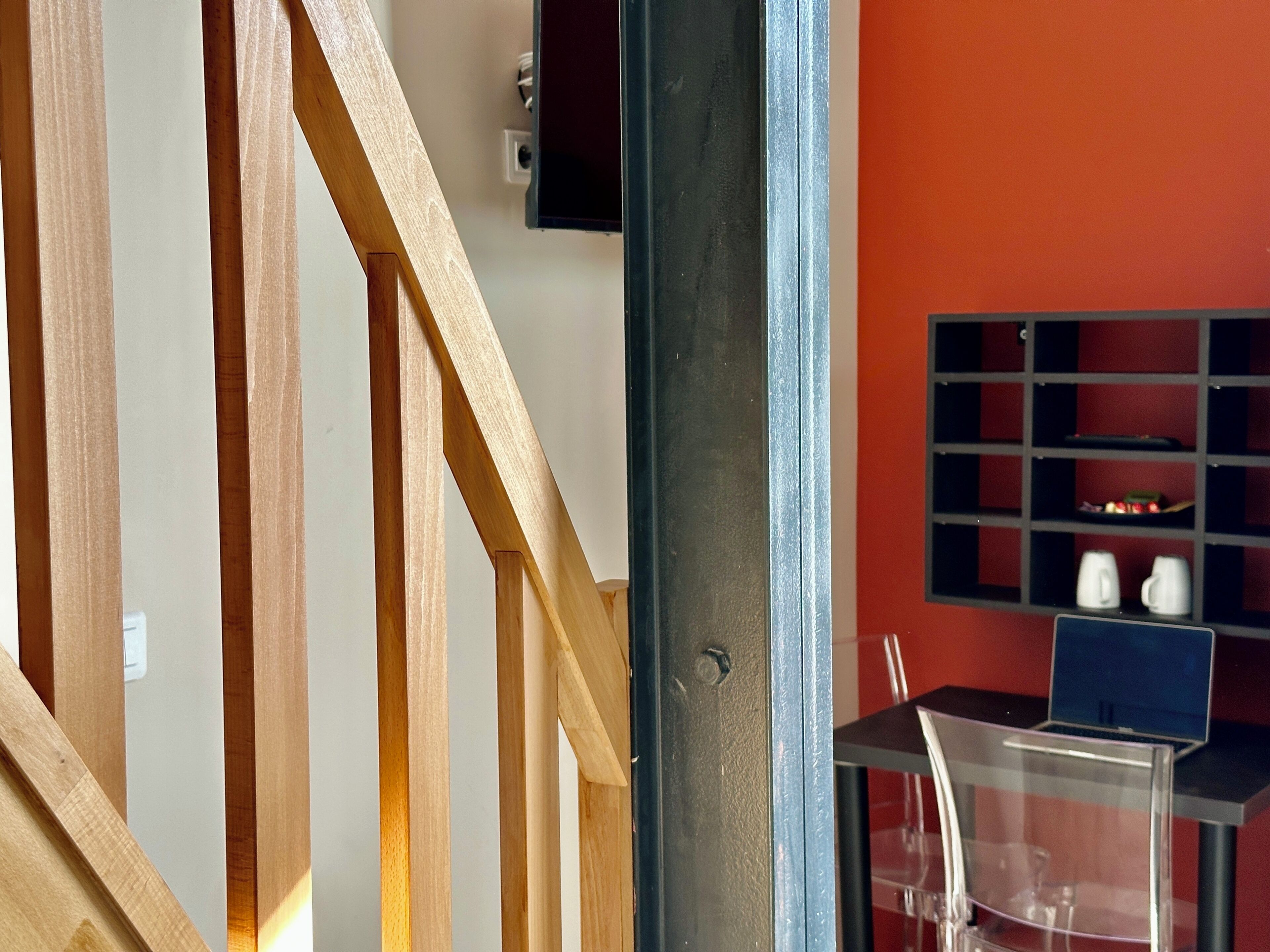Una escalera con barandillas de madera, un marco de puerta metálico y una pared roja con un estante negro que sostiene tazas y una computadora portátil.