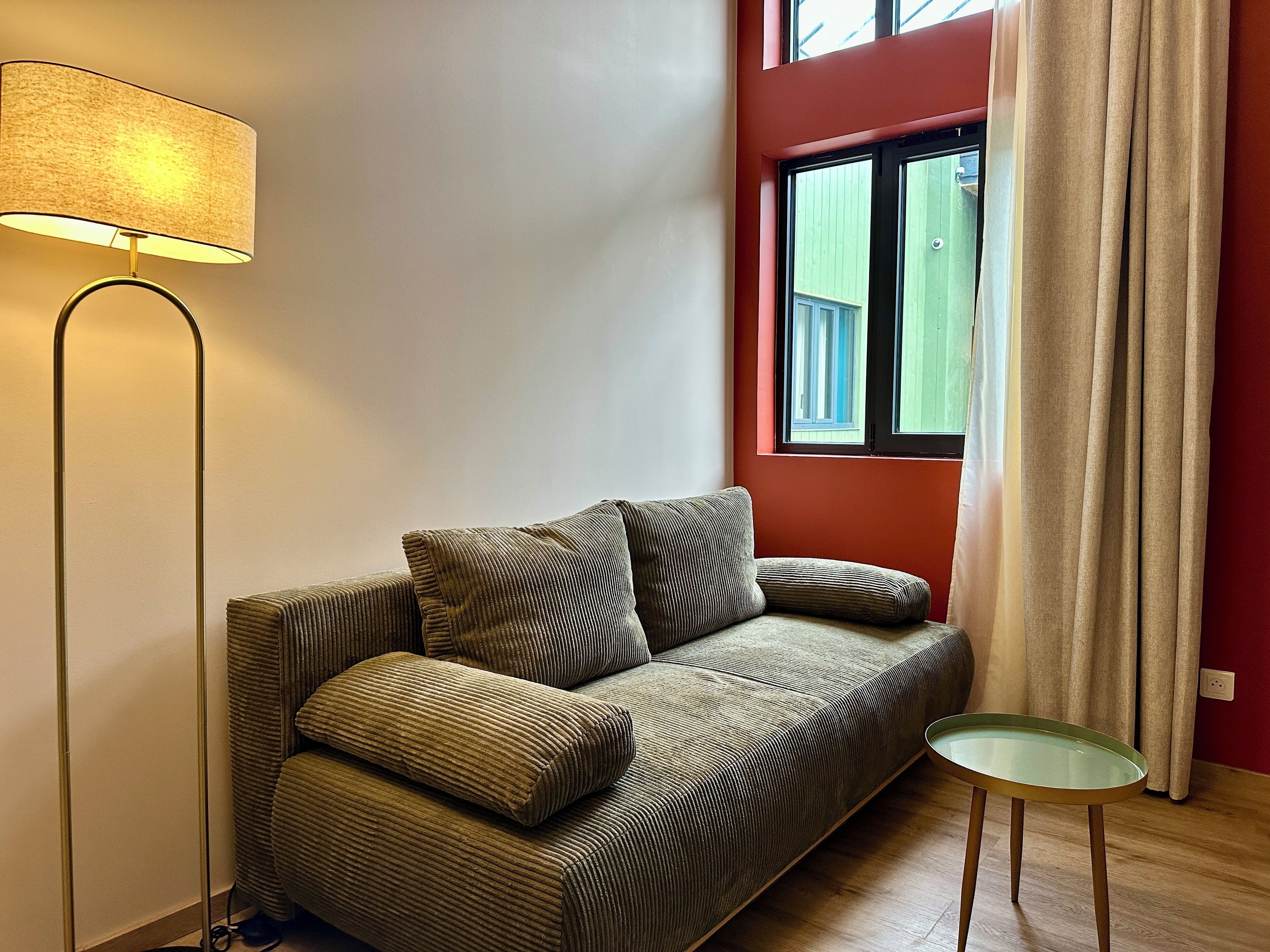 Een woonkamer met een bank, een vloerlamp, een bijzettafeltje en een raam met gordijnen.