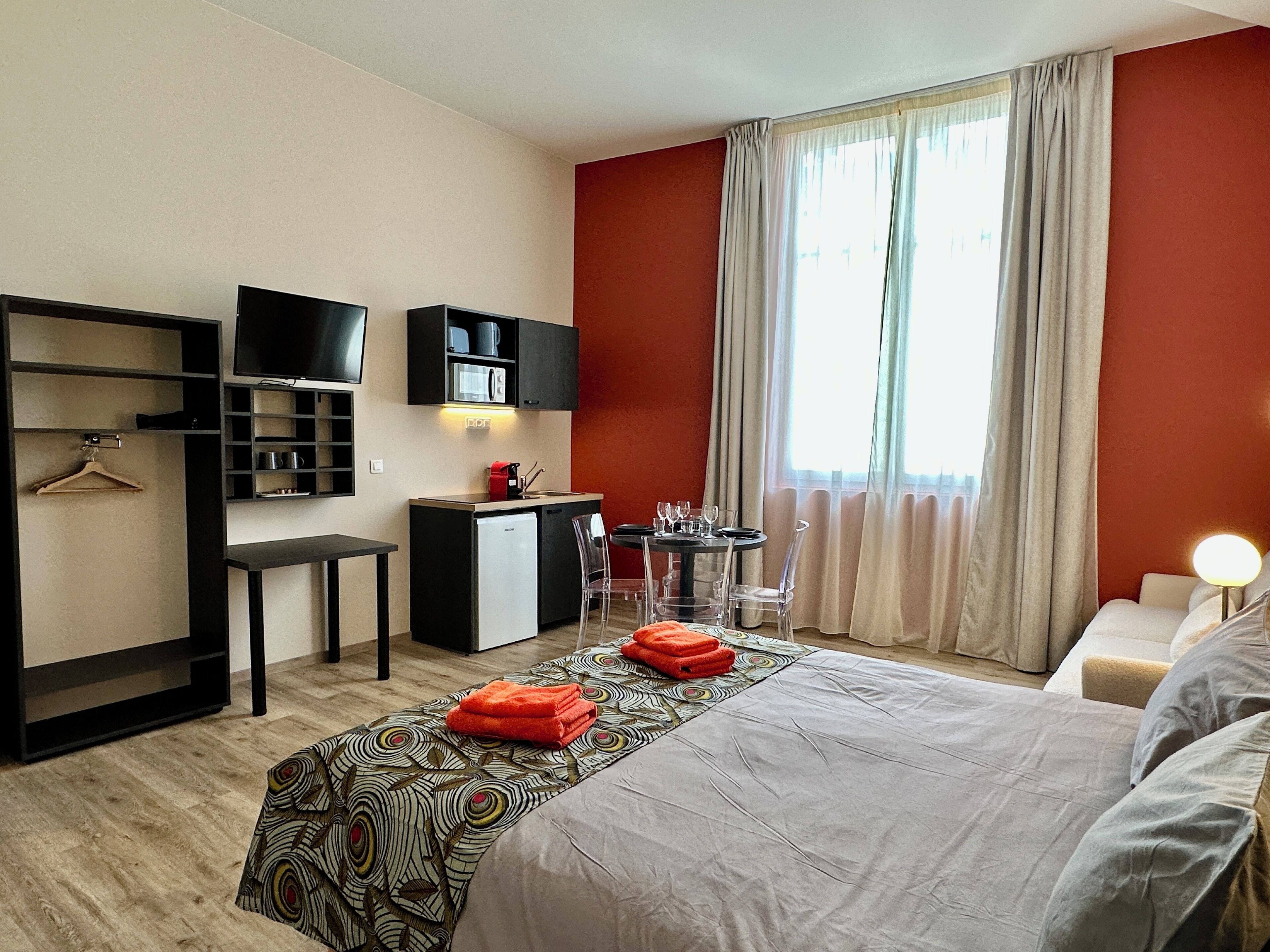 A hotel room with a bed, a desk with a computer, a small kitchenette, and a TV.