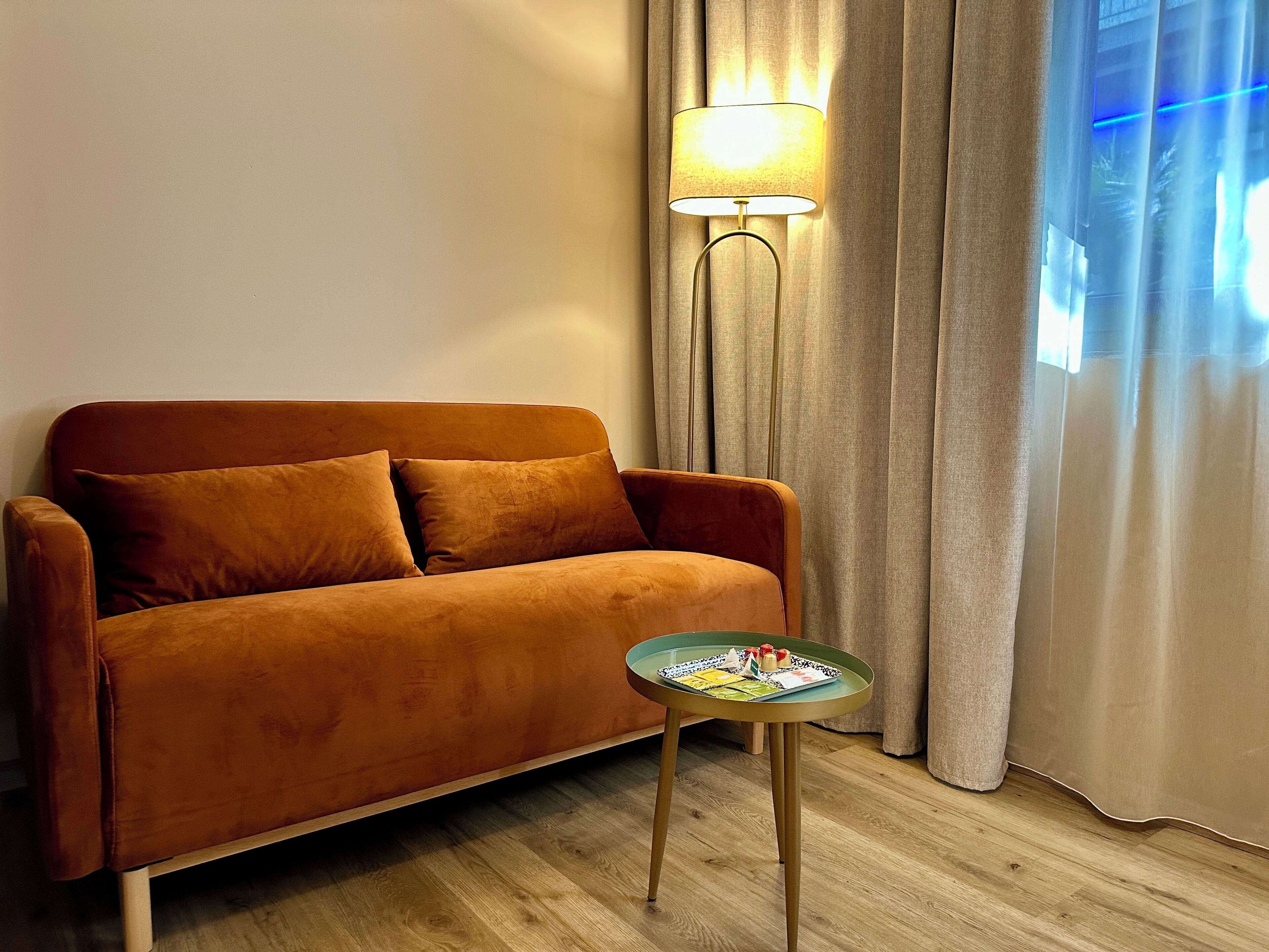 Ein braunes Sofa mit zwei Kissen, ein kleiner runder Tisch mit einer Snackplatte, eine Stehlampe und ein Fenster mit Vorhängen.