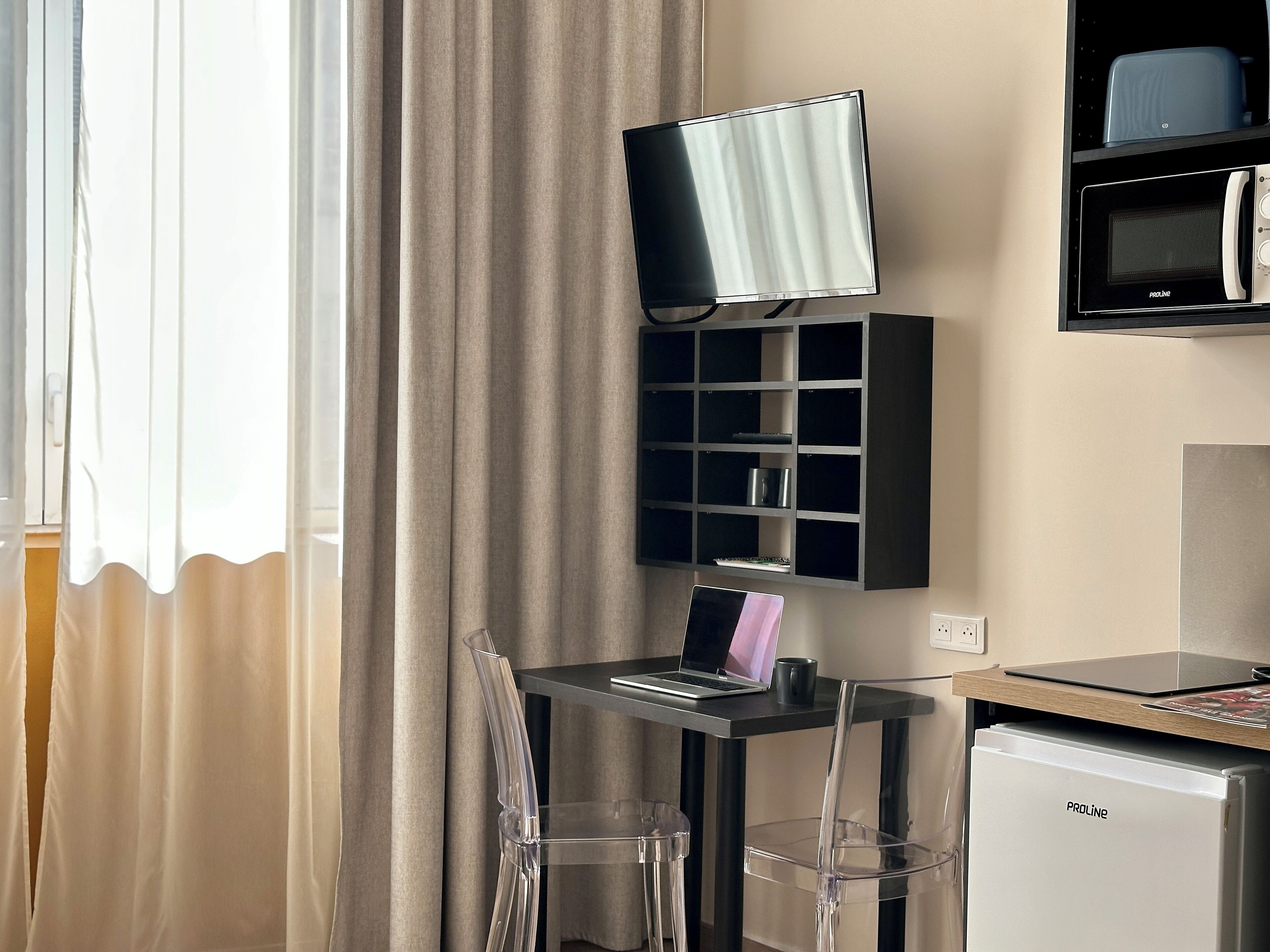 A hotel room with a desk, a flat-screen TV mounted on the wall, a mini-fridge, and a microwave.