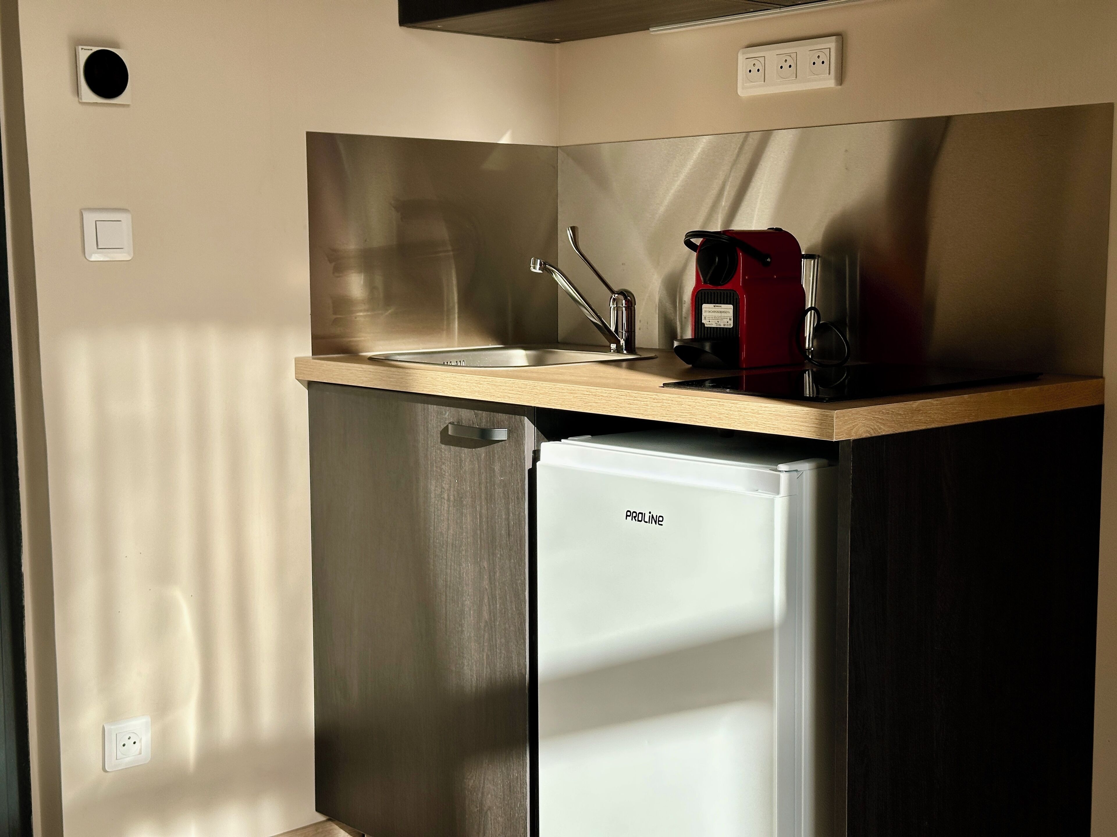 A compact kitchenette with a stainless steel refrigerator, a sink, and a red coffee maker on the countertop.
