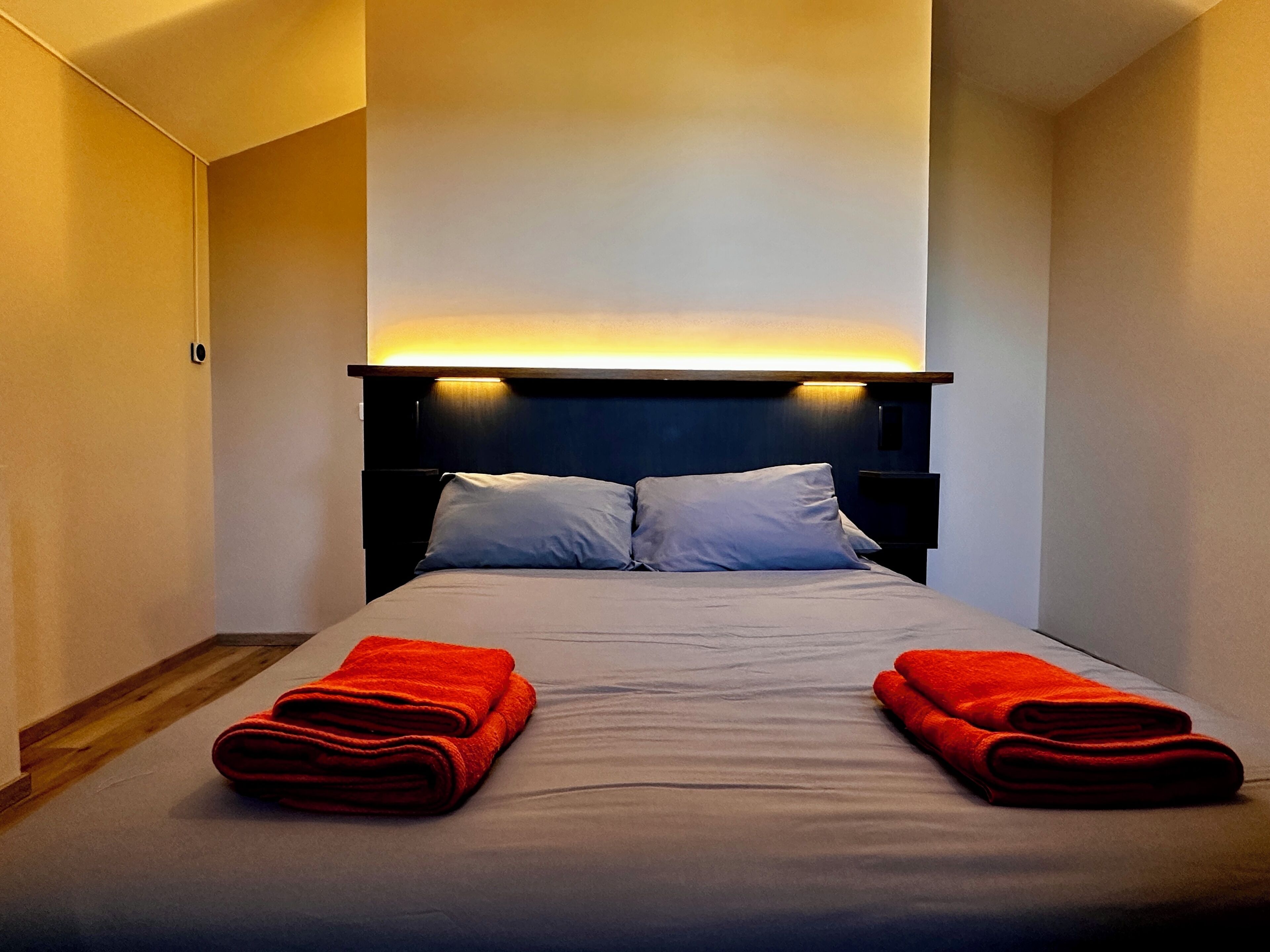 Habitación de hotel con una cama, dos almohadas, cabecera con iluminación integrada y dos toallas rojas sobre la cama.