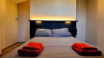 Habitación de hotel con una cama, dos almohadas, cabecera con iluminación integrada y dos toallas rojas sobre la cama.