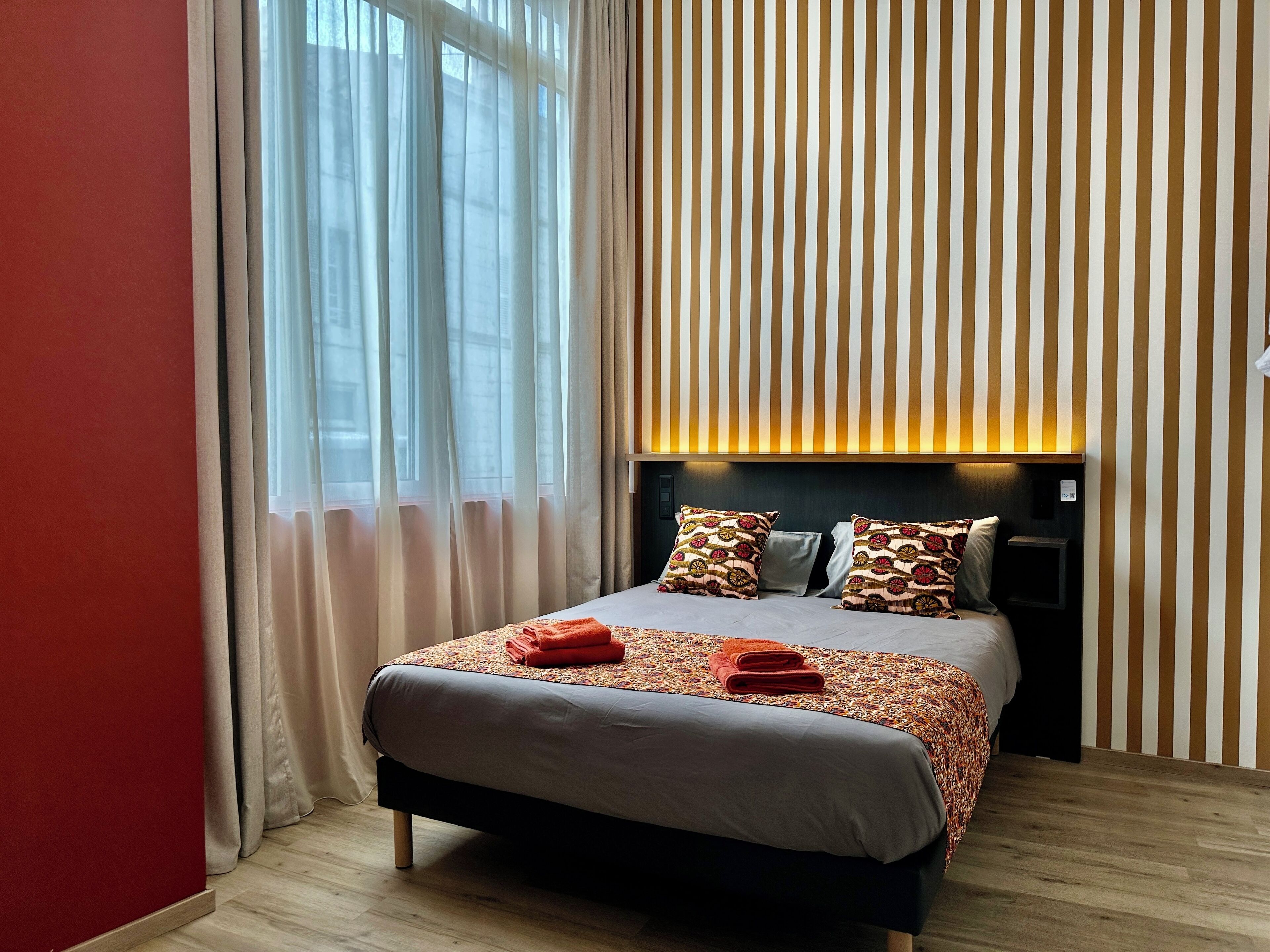 Une chambre d’hôtel avec un lit, des rideaux, un mur à rayures et une table de chevet.