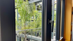 Vista desde un balcón con barandillas metálicas y piso a cuadros, con vista a un edificio con vegetación y un banco rojo.