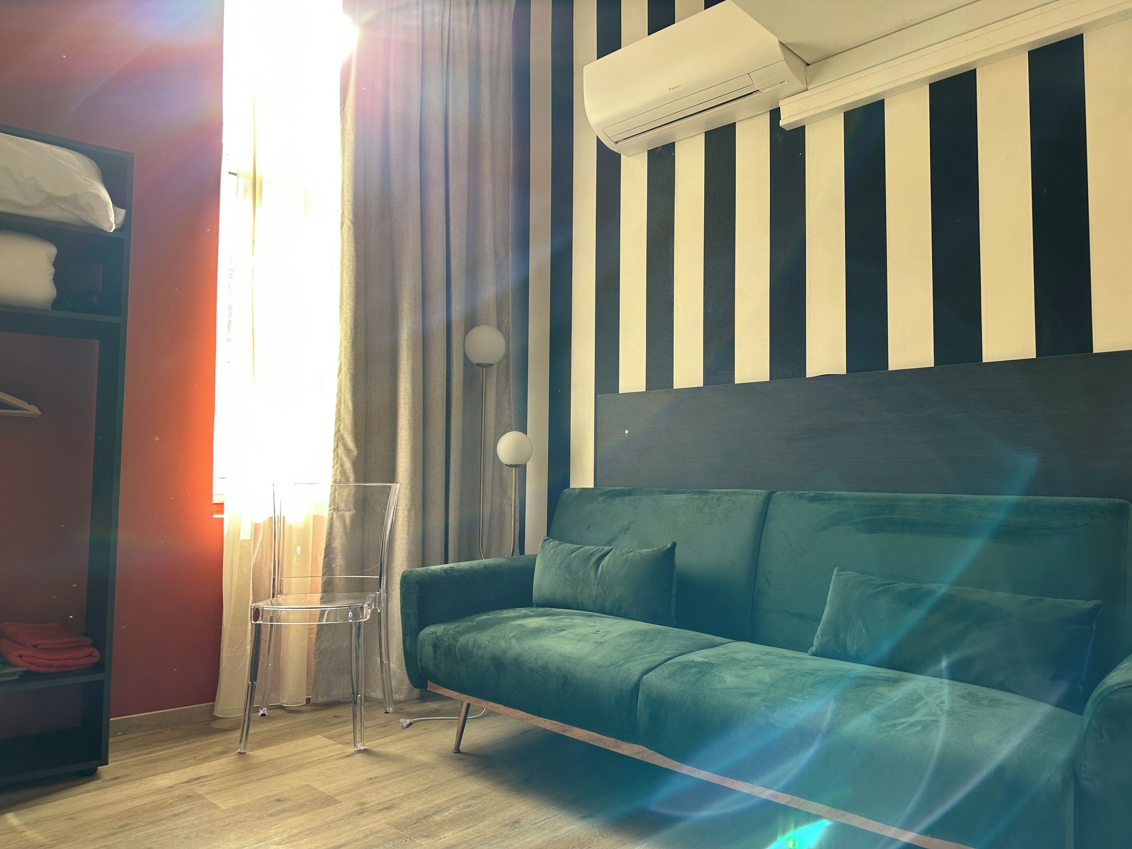 A room with a green sofa, a bed with a white headboard, a window with curtains, an air conditioning unit, and a striped wall.