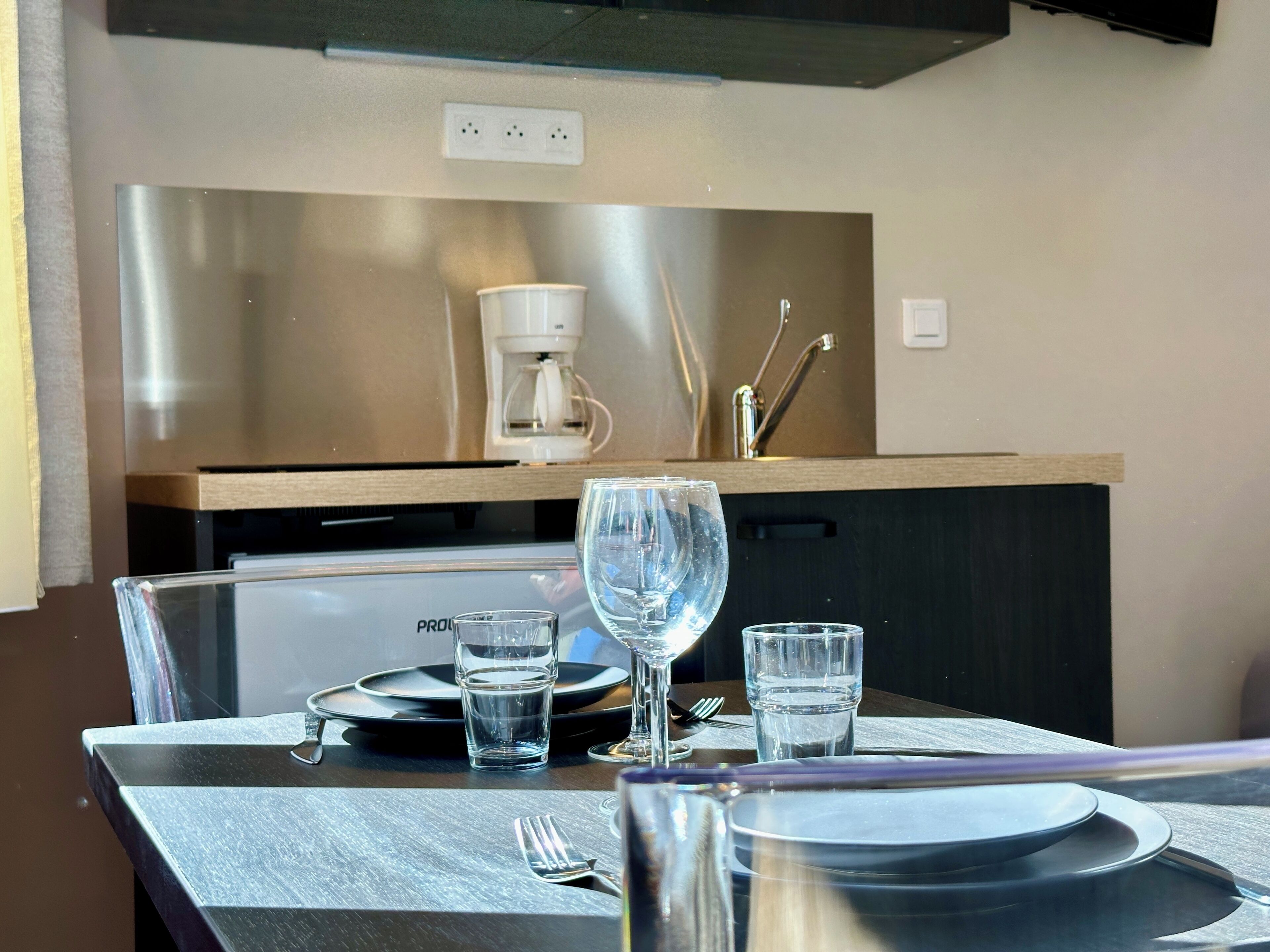 Una cocina moderna con electrodomésticos de acero inoxidable, una cafetera y un comedor con una mesa preparada para dos personas, incluyendo platos, vasos y cubiertos.
