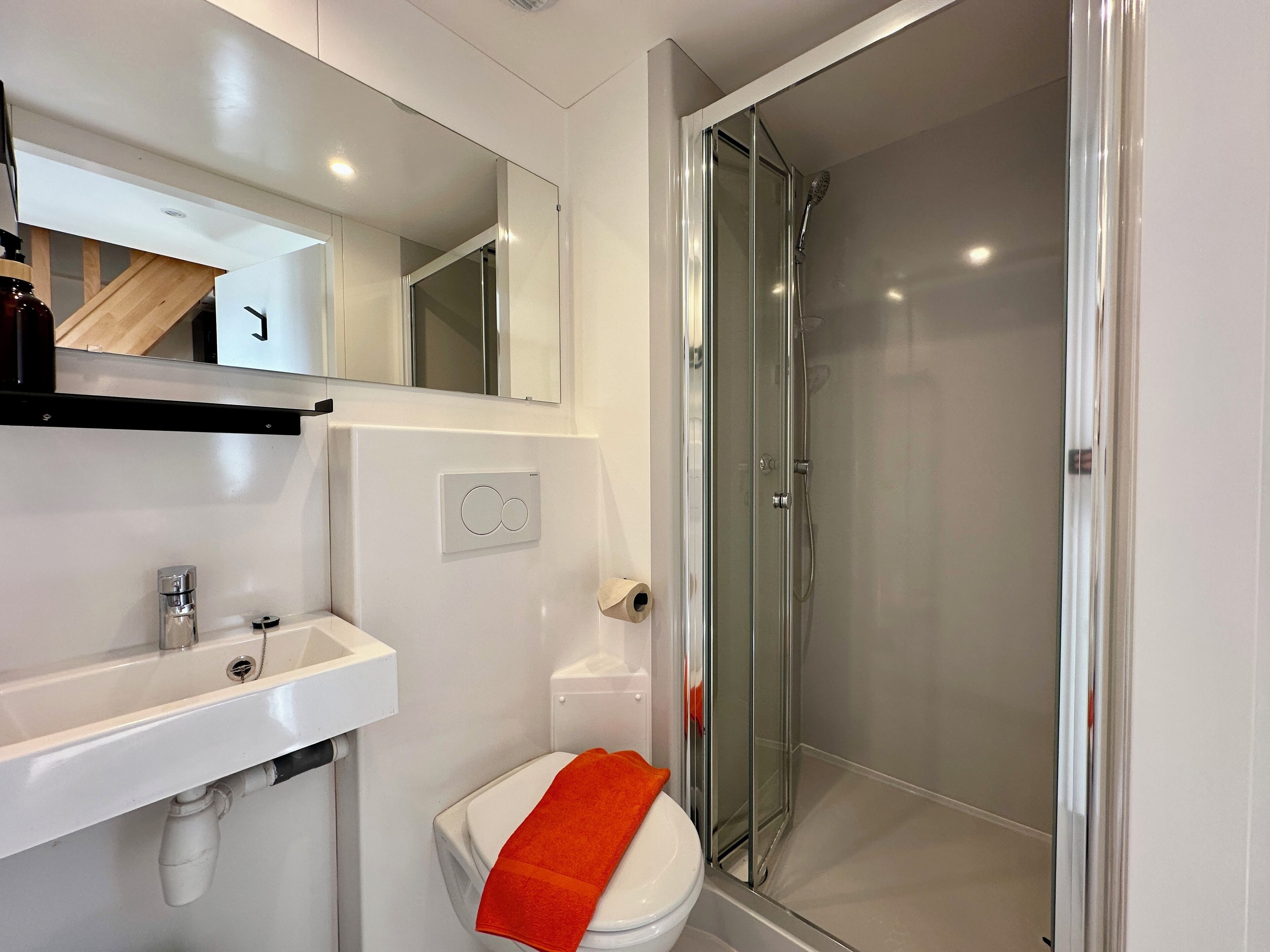 Un baño moderno con ducha de vidrio, un lavamanos blanco y un inodoro con una toalla naranja.