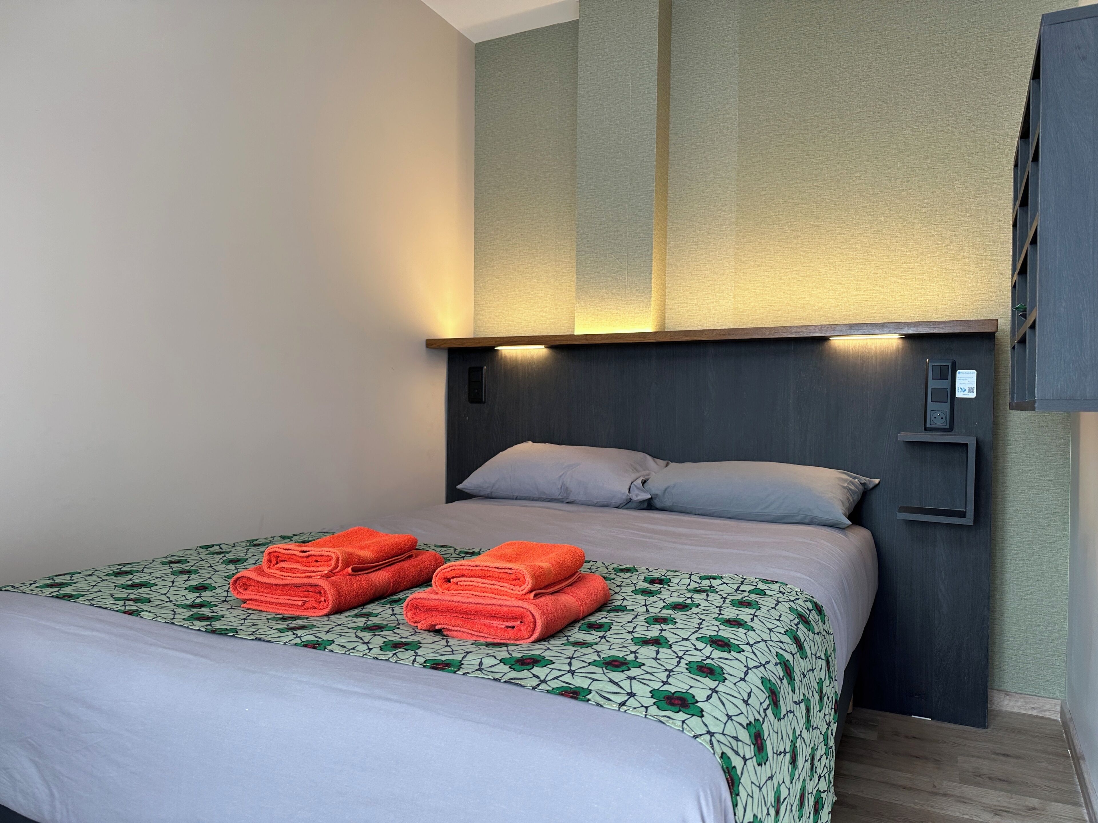 A hotel room with a neatly made bed, a headboard with built-in lighting, and a wall-mounted control panel.