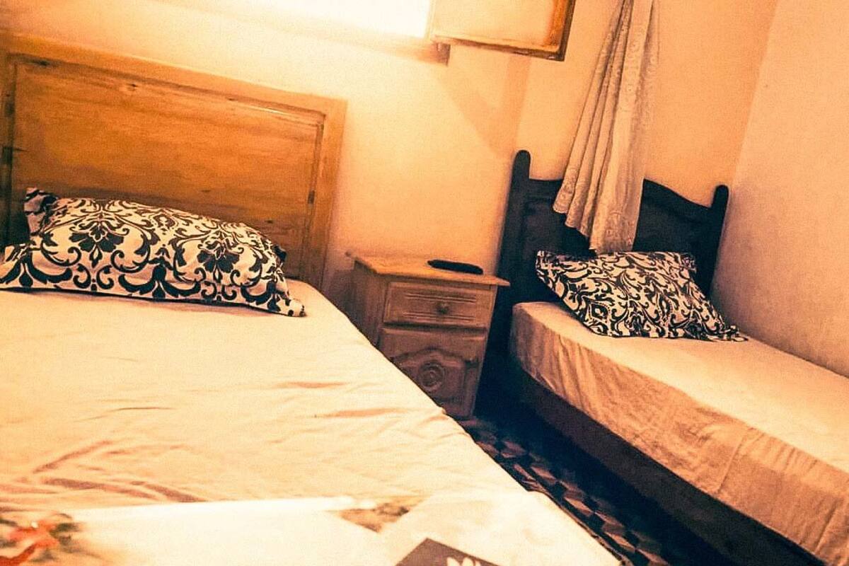 Et værelse med to senge, hver med mønstrede puder og en trægavl. Der er et natbord med en lampe og en lille skuffe.
