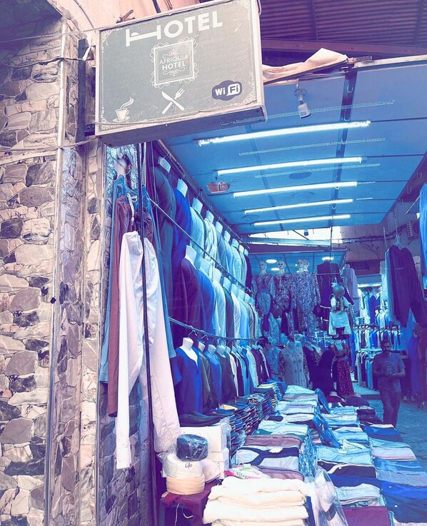 A narrow corridor with clothing racks on the left and a variety of folded clothes on the right. A sign above indicates it's a hotel. - Hotel Afriquia (Oujda)