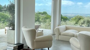 A spacious room with large windows offering a view of a lush landscape. There are two white armchairs and a small table.