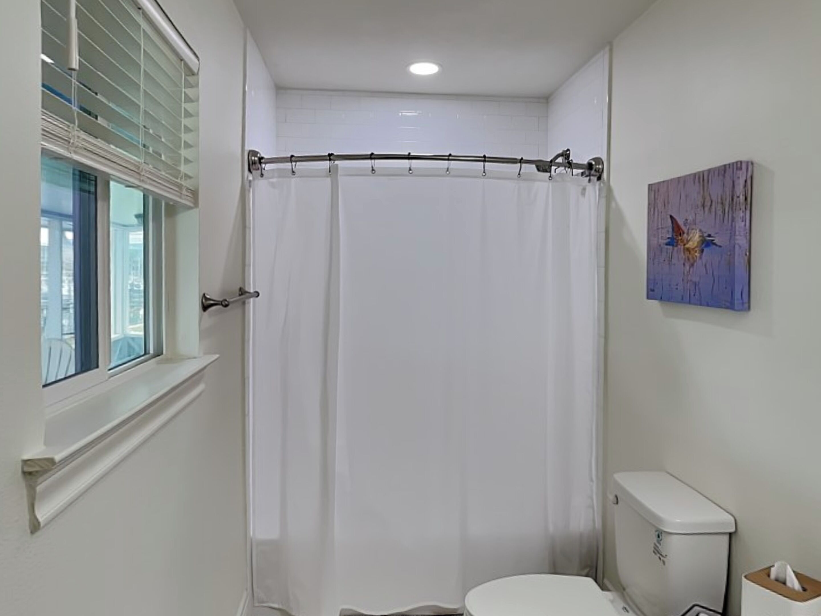 A small, clean bathroom with a shower curtain, toilet, and a window with blinds.